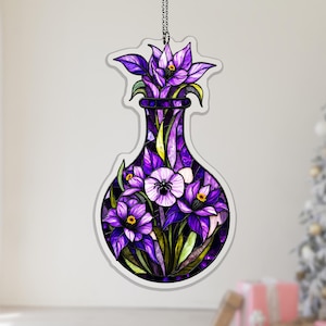 May include: A decorative ornament featuring a stained-glass style design of a vase filled with purple flowers. The flowers have yellow centers and green leaves. The vase is a deep purple color and is hanging from a silver chain.