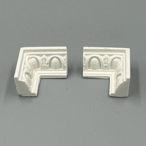 May include: Two white corner moldings with ornate designs. The moldings are made of a lightweight material and have a smooth finish.