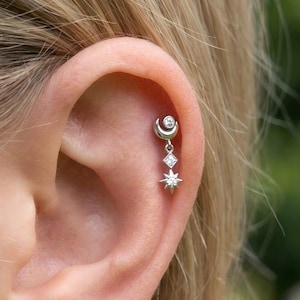 May include: A silver earring featuring a crescent moon, a diamond-shaped gem, and a star pendant. The earring is dangling from the ear. The design is delicate and celestial, suitable for cartilage piercings.