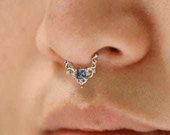 Puede incluir: Un anillo de septum plateado con una gema azul cuadrada en el centro. El diseño ornamentado presenta delicados adornos y pequeñas piedras de acento transparentes. La joya se lleva en la nariz de una persona.