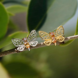 Könnte beinhalten: Zwei schmetterlingsförmige Ohrringe auf einem dünnen Zweig. Ein Ohrring ist silberfarben mit filigranem Flügelmuster und orangefarbenen Steinen, der andere ist goldfarben mit passendem filigranem Flügeldesign und orangefarbenen Steinen.
