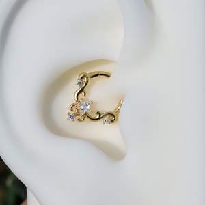 May include: A gold-colored septum ring with a decorative design. The ring features a star-shaped gem and smaller gems, with a swirling pattern. The jewelry is shown on a white background.
