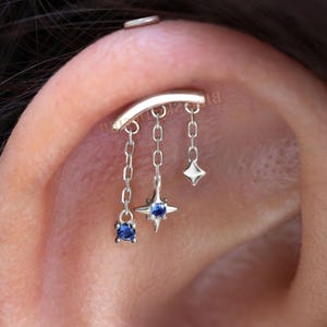 Può includere: Un piercing all'orecchio in argento con una barra curva e tre ciondoli pendenti. I ciondoli includono una gemma blu quadrata, un design a stella con un centro di gemma blu e un ciondolo a forma di diamante. Il piercing è mostrato su un orecchio.