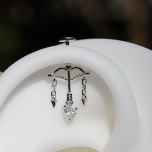 Puede incluir: Una joya de piercing corporal plateada con un dise&ntilde;o de arco y flecha con cadenas y puntas de flecha colgantes. Una gema transparente en forma de coraz&oacute;n cuelga del centro. La joya est&aacute; sobre un fondo blanco.