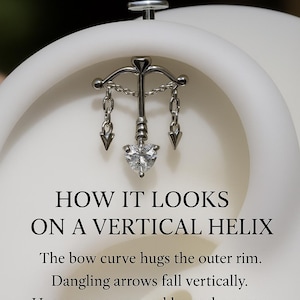Puede incluir: Un pendiente vertical de h&eacute;lice plateado con un dise&ntilde;o de arco, flechas colgantes y una piedra en forma de coraz&oacute;n. El texto dice: "HOW IT LOOKS ON A VERTICAL HELIX". La curva del arco abraza el borde exterior. Las flechas colgantes caen verticalmente. La piedra en forma de coraz&oacute;n brilla en el centro. Elegante y dram&aacute;tico.