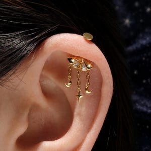 May include: A gold-toned cartilage earring featuring a moth-shaped top adorned with small clear stones. Three delicate chains hang from the moth, each ending in a charm: two crescent moons on the outer sides and a four-pointed star in the center.