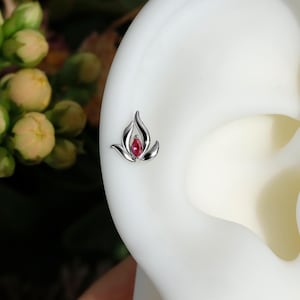 May include: A silver-colored stud earring in the shape of a stylized flame, with a small, oval-shaped, red gemstone at its center. The earring is set against a white background, with a blurred green and yellow floral element in the background.