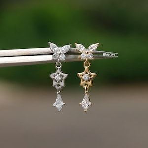 Può includere: Orecchini con design a farfalla, stella e pendente a forma di diamante. Un orecchino è argentato, l'altro dorato. Il testo "Blue Sky" è visibile. Sfondo verde sfocato.