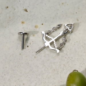 Puede incluir: Un pendiente de plata y un pendiente a juego con un dise&ntilde;o de arco y flecha con una cadena. El pendiente tiene un detalle en forma de coraz&oacute;n en el extremo de la flecha. La joyer&iacute;a se exhibe sobre una superficie clara y moteada.