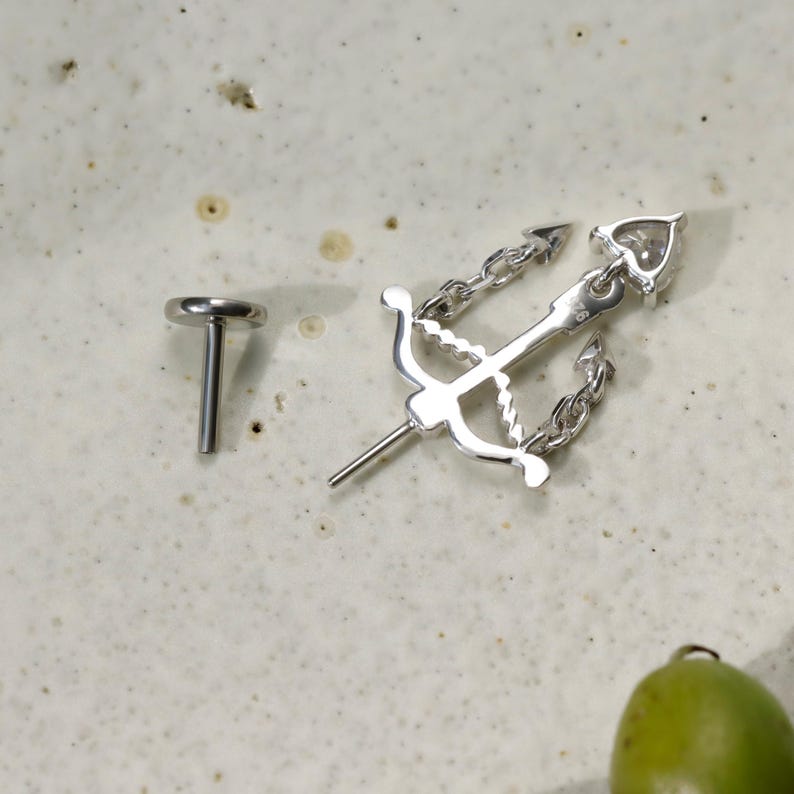 Puede incluir: Un pendiente de plata y un pendiente a juego con un dise&ntilde;o de arco y flecha con una cadena. El pendiente tiene un detalle en forma de coraz&oacute;n en el extremo de la flecha. La joyer&iacute;a se exhibe sobre una superficie clara y moteada.
