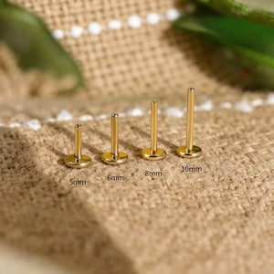 Może przedstawiać: Cztery złote kolczyki do piercingu warg o różnych rozmiarach: 5 mm, 6 mm, 8 mm i 10 mm. Każdy kolczyk ma płaską, okrągłą podstawę i prosty pręt. Kolczyki są prezentowane na teksturowanej, beżowej powierzchni.