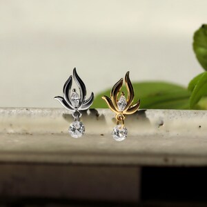 May include: Two decorative charms, one silver-toned and one gold-toned, each featuring a flame-like metal frame with a central marquise-cut clear stone and a dangling round clear stone at the base, designed for attachment to jewelry or accessories.