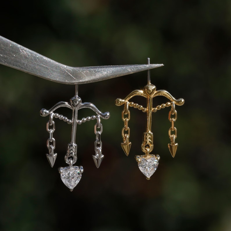 Puede incluir: Primer plano de dos pendientes, uno plateado y otro dorado. Cada pendiente presenta un dise&ntilde;o de arco y flecha con cadenas colgantes y una gema en forma de coraz&oacute;n. Los pendientes son sostenidos por una herramienta plateada sobre un fondo verde borroso.