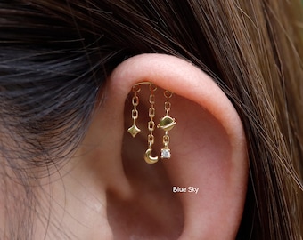 Peut inclure: Bijou d'oreille doré sur le thème céleste. Le design comprend un charm en forme de losange, un croissant de lune, une planète et un petit diamant, tous suspendus par de fines chaînes. Le texte "Blue Sky" est visible.