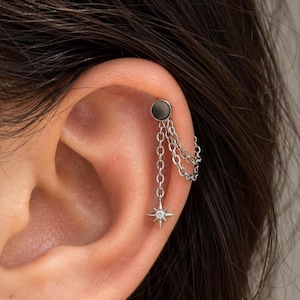 Può includere: Un orecchino da cartilagine color argento con un perno circolare collegato a due catenelle sottili. Una catena termina con un piccolo ciondolo a forma di stella a quattro punte, impreziosito da una pietra chiara e brillante al centro.