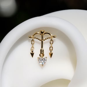 May include: A gold-colored earring featuring a bow and arrow design with a heart-shaped gemstone. The earring has a delicate chain and arrow pendants. The gemstone is clear and faceted, reflecting light.