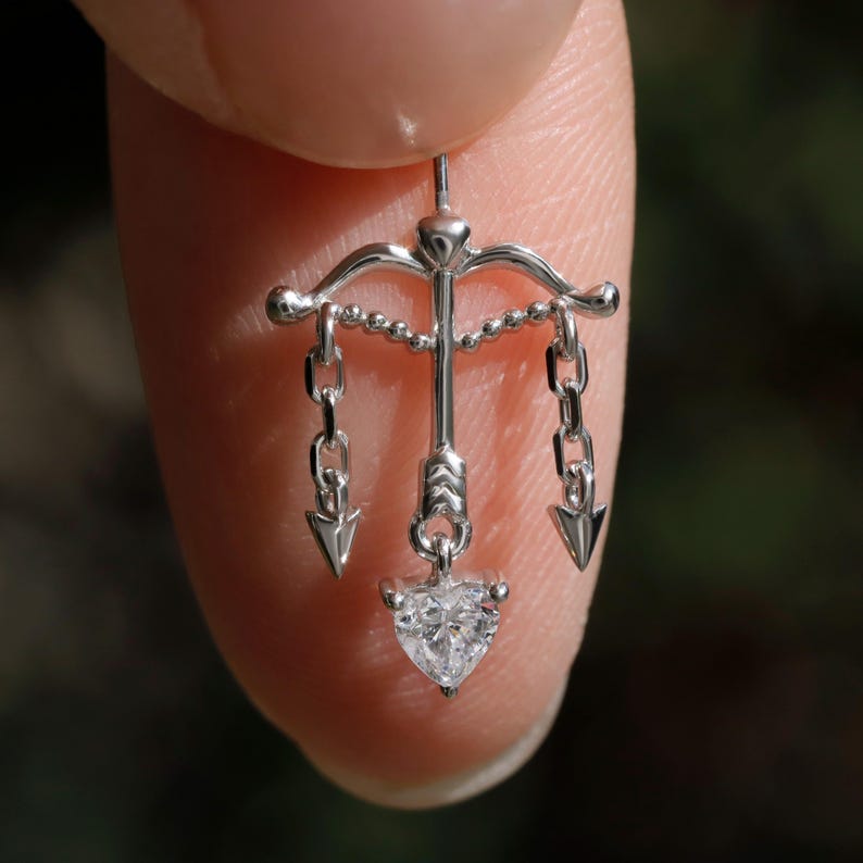Puede incluir: Un peque&ntilde;o colgante plateado con forma de arco y flecha, con un diamante en forma de coraz&oacute;n en la parte inferior. El colgante tiene tres flechas colgando de cadenas y se sostiene entre dos dedos.