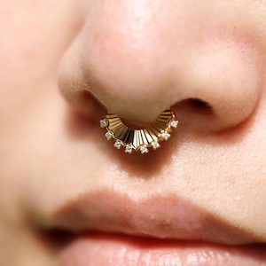 Puede incluir: Primer plano de un aro de septum dorado con diseño en forma de abanico. El piercing nasal presenta pequeñas gemas transparentes. La imagen destaca los detalles del piercing y la joya dorada.