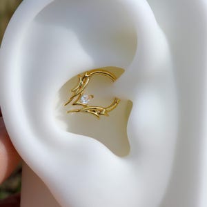 May include: A gold-colored, hinged earring with a decorative, stylized design. The earring features a small, clear gemstone at its center. The design includes curved lines and pointed elements, creating a unique and elegant look. The earring is shown in an ear piercing.