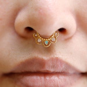 May include: A gold septum ring with a scalloped design. The ring features small clear stones and a central opal-like stone. The design is accented with small gold beads. The text "Blue Sky" is visible in the image.