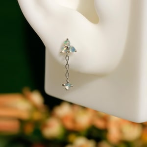 May include: A silver earring with a cluster of three opalescent stones at the top, connected by a delicate chain to a single, square-shaped opalescent stone. The earring is displayed on a white surface.