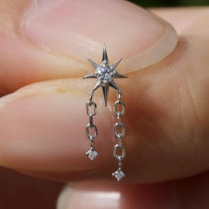 以下が含まれることがあります： 星型のデザインのシルバーイヤリング。中央にダイヤモンドがあり、チェーンが垂れ下がっています。各チェーンの端には小さなダイヤモンドが付いています。イヤリングは片耳のピアス用に設計されています。