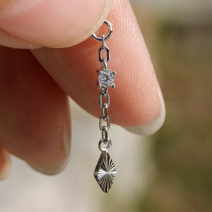 May include: A silver earring with a small, clear gemstone and a diamond-shaped charm. The earring is suspended from a chain and is held up by a finger.