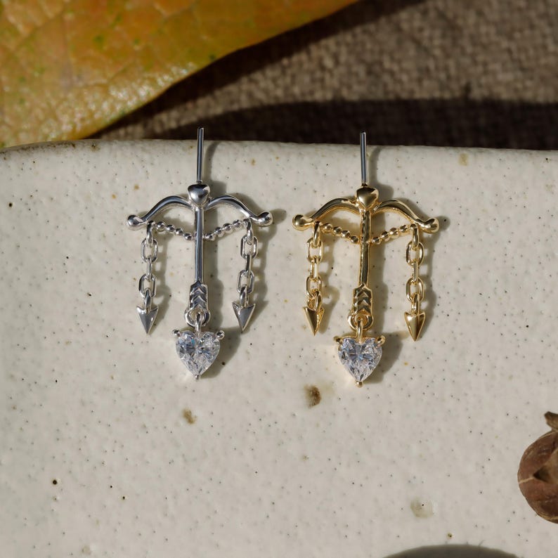 Puede incluir: Dos pendientes, uno plateado y otro dorado, cada uno con un dise&ntilde;o de arco y flecha. Los pendientes tienen una gema en forma de coraz&oacute;n en la parte inferior y detalles de cadena colgantes. Se muestran sobre una superficie de tono neutro.