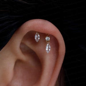 May include: Two gold-toned cartilage earrings are worn in the upper ear. One earring features a single marquise-cut clear stone. The second earring consists of a small round clear stone stud with a dangling marquise-cut clear stone charm.