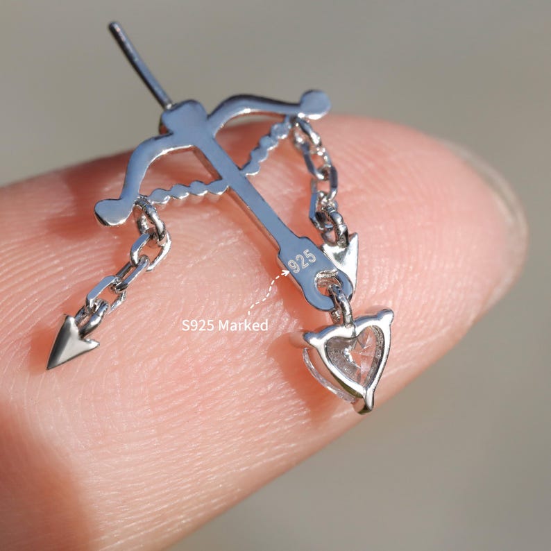 Puede incluir: Un colgante plateado con forma de arco y flecha, con una punta de flecha en forma de coraz&oacute;n. El colgante est&aacute; marcado con "925" y conectado a una cadena. El texto "S925 Marked" tambi&eacute;n es visible.
