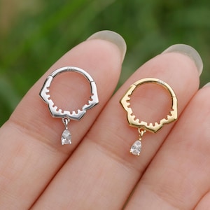 May include: Two earrings, one silver and one gold, each featuring a floral-shaped hoop with a teardrop-shaped gemstone pendant. The earrings are displayed against a blurred green background.