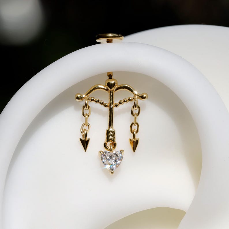 Puede incluir: Pendiente dorado con dise&ntilde;o de arco y flecha, con una piedra transparente en forma de coraz&oacute;n. El pendiente tiene un poste para insertar y est&aacute; sobre un fondo blanco.