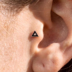 Könnte beinhalten: Ein kleiner dreieckiger Ohrstecker mit einer schwarzen Emaileinlage, die von einem perlenartigen silberfarbenen Rand umgeben ist, und einem einzelnen klaren runden Edelstein in der Mitte. Der Ohrstecker sitzt am Tragus des Ohrs.