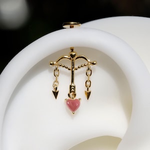 May include: A gold-colored earring featuring a bow and arrow design with a pink heart pendant. The earring has a post backing and is set against a white background. The arrowheads and bow are detailed.