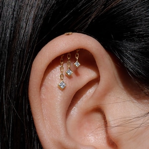 May include: Close-up of a gold ear piercing with three dangling chain earrings, each featuring a small, square, clear gemstone. A small, round gold stud is also visible at the top of the ear. The jewelry is set against the backdrop of dark hair and skin.