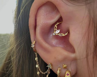 May include: Gold-toned jewelry set on an ear, including a septum ring with a marquise-cut stone and a chain earring. Other earrings include a small hoop, a star stud, and a hoop with colorful stones. The jewelry is delicate and stylish.