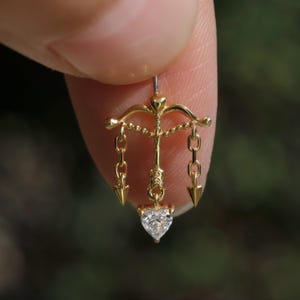Puede incluir: Un pendiente de color dorado con una gema en forma de coraz&oacute;n en la parte inferior. El pendiente presenta un dise&ntilde;o de arco y flecha con cadenas y puntas de flecha colgantes. El pendiente se sostiene entre dos dedos.