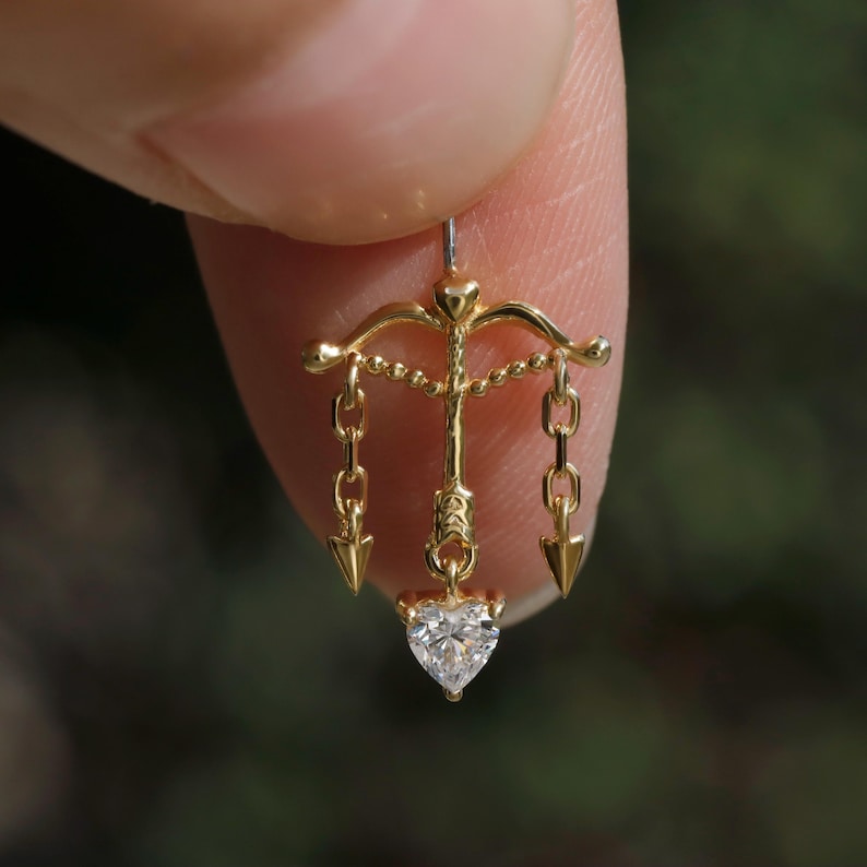 Puede incluir: Un pendiente de color dorado con una gema en forma de coraz&oacute;n en la parte inferior. El pendiente presenta un dise&ntilde;o de arco y flecha con cadenas y puntas de flecha colgantes. El pendiente se sostiene entre dos dedos.