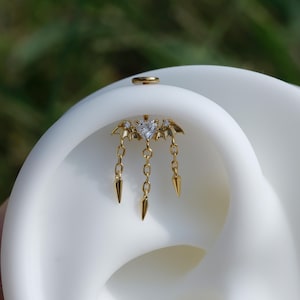 May include: A gold ear piercing with a heart-shaped gem and dangling chain details. The jewellery has a bat-wing design with three chains ending in pointed shapes. A small gold ring is also visible.