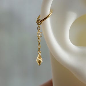 May include: A gold earring featuring a chain with a diamond-shaped charm. The earring is attached to a hoop. The charm has a textured, sunburst design. The earring is clipped to a white ear.