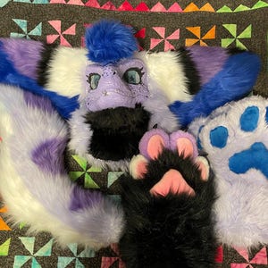 以下が含まれることがあります： 紫と青のドラゴンの頭、尻尾、足パッドを備えた毛皮のコスチュームセット。頭には青い毛皮の房と詳細な顔の特徴があります。尻尾は白、紫、青の毛皮のミックスです。