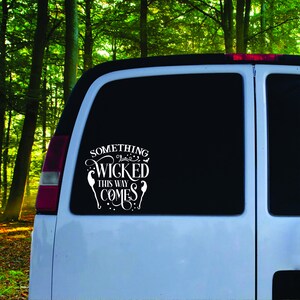 Puede incluir: Una calcomanía de vinilo blanco en una ventana negra de furgoneta. La calcomanía dice "SOMETHING WICKED THIS WAY COMES" en una fuente decorativa, con acentos de bruja. El fondo es un bosque.