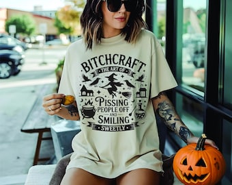 Könnte beinhalten: Cremefarbenes T-Shirt mit schwarzem Aufdruck: "Bitchcraft: The Art of Pissing People Off and Smiling Sweetly." Das Design zeigt eine Hexe, Krähen, einen Kessel und eine Zaubertrankflasche. Eine geschnitzte Kürbis wird gehalten.