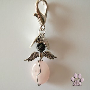 May include: A silver-toned angel charm with a lobster clasp. The charm features a pink oval gemstone body, a black bead head, and detailed silver wings. Small silver accents and charms hang above the wings.