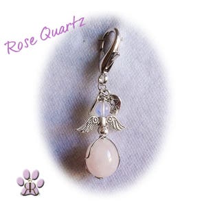 May include: A silver-toned charm with a lobster clasp, featuring an angel design with wings, a heart, and a rose quartz gemstone. The text "Rose Quartz" is written in a cursive font. A paw print logo is in the lower left corner.