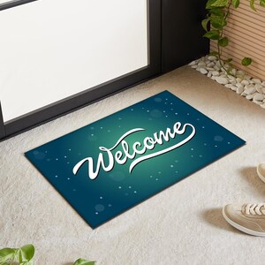Puede incluir: Una alfombra de bienvenida azul verdosa con texto blanco que dice "Welcome" en una fuente cursiva. La alfombra tiene un efecto de brillo sutil.