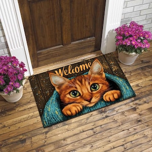 Könnte beinhalten: Eine rechteckige Fußmatte mit einem Katzendesign. Die Matte zeigt eine große orangefarbene Tabby-Katze mit grünen Augen, die aus einer blauen Strickdecke lugt. Das Wort "Welcome" ist in goldenen Buchstaben aufgedruckt.
