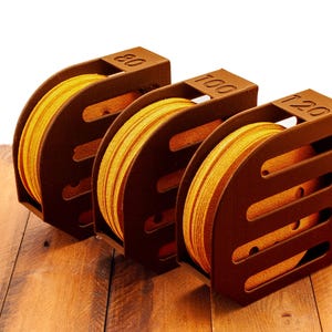 Puede incluir: Tres cajas de almacenamiento de madera marr&oacute;n para discos de lija. Las cajas est&aacute;n etiquetadas con 80, 100 y 120. Cada caja contiene un rollo de discos de lija amarillos.