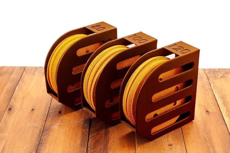 Puede incluir: Tres cajas de almacenamiento de madera marr&oacute;n para discos de lija. Las cajas est&aacute;n etiquetadas con 80, 100 y 120. Cada caja contiene un rollo de discos de lija amarillos.