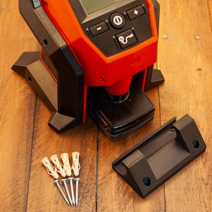 May include: A red and black power tool with a digital display and a black base. The tool is sitting on a wooden surface. There are four white plastic anchors and four silver screws next to the tool.